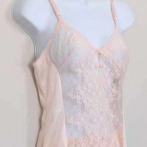 Op de afbeelding: Zachtroze vintage onderjurk met delicate kanten details. De bodice heeft een doorschijnend kanten paneel met bloempatronen en de rok is een vloeiende, effen stof. De bandjes zijn dun en verstelbaar en de zoom is afgezet met kant.