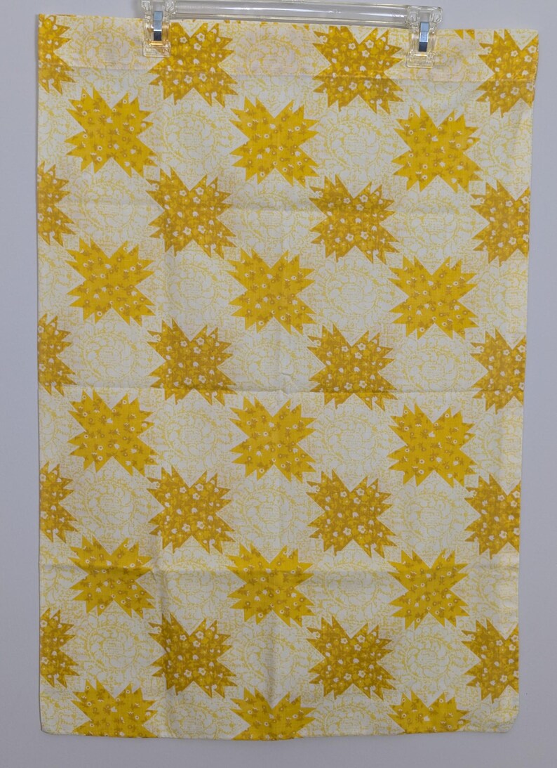K&ouml;nnte beinhalten: Ein rechteckiger, cremefarbener Stoff mit einem sich wiederholenden Muster aus gelben Sternen. Jeder Stern ist mit kleinen wei&szlig;en floralen Details gef&uuml;llt. Der Stoff h&auml;ngt an einem transparenten Kunststoffclip.