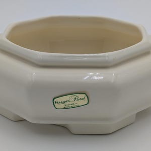 May include: Cream-colored ceramic Haeger Floral planter with an octagonal shape. The planter has a wide rim and a slightly raised base. A green and white label is affixed to the front.