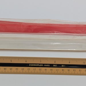 Puede incluir: Una regla de madera con marcas negras y una curva de plástico roja en una funda de plástico transparente. La regla está marcada en centímetros. La curva roja está en una funda transparente.