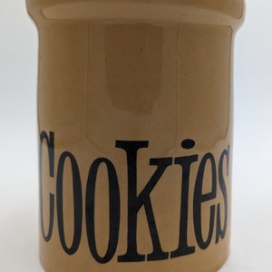 Puede incluir: Tarro de galletas de cerámica de color beige con la palabra "COOKIES" en letras negras grandes. El tarro tiene una parte superior redondeada y una forma cilíndrica. Un recipiente de cocina clásico.