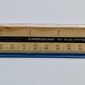 Puede incluir: Una regla de madera con marcas negras y un asa de plástico azul. La regla está marcada en centímetros y tiene una funda de plástico transparente. El texto "DUNLOP MADE IN JAPAN" es visible.