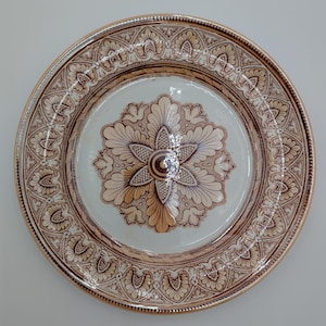 Puede incluir: Un plato de cerámica decorativo con un diseño floral. El plato presenta un motivo floral central en tonos marrón y beige, rodeado por un borde detallado. El borde tiene un ribete dorado. El plato se utiliza probablemente para servir o exhibir, con un diámetro de unos 25 cm.