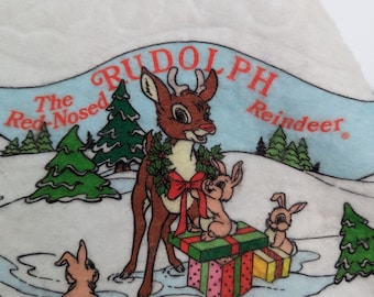 Op de afbeelding: Een witte kerstsok van vilt met een Rudolph the Red-Nosed Reindeer-ontwerp. Rudolph is afgebeeld in een besneeuwde scène met een rode strik en een krans om zijn nek. Hij houdt een stapel ingepakte cadeaus vast met drie konijntjes in de buurt. De sok heeft de tekst "Rudolph The Red-Nosed Reindeer" erop gedrukt.