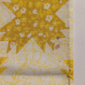 K&ouml;nnte beinhalten: Nahaufnahme eines Stoffabschnitts mit einem sich wiederholenden Sternmuster. Die Sternmuster sind gelb und mit winzigen wei&szlig;en Blumen gef&uuml;llt. Der Hintergrund ist hellgelb mit einem dezenten Blumendesign. Es gibt ein paar kleine braune Flecken auf dem Stoff.