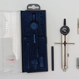 May include: A drawing compass set featuring a silver and black compass, spare parts, and a black case with a blue velvet interior. The set includes a clear plastic lid, designed for precise drawing and measurement.