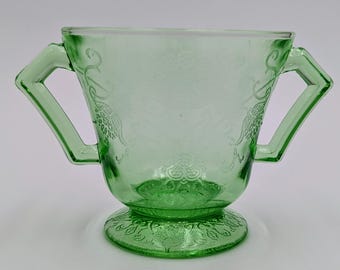 May include: A green glass sugar bowl with two handles and a footed base. The bowl has an embossed floral design.