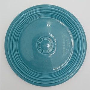 Puede incluir: Una tapa de cerámica de color turquesa con detalles concéntricos acanalados. La tapa tiene un pomo central y un acabado brillante. El diseño circular sugiere que es para un cuenco o una olla, probablemente para uso en la cocina o el comedor.