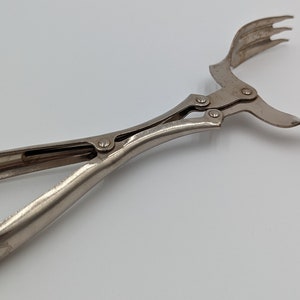 Op de afbeelding: Zilverkleurige metalen tang met een scharnierend handvat en een drietandvormig grijpend uiteinde. De tang is ontworpen om kleine voorwerpen op te pakken en vast te houden.