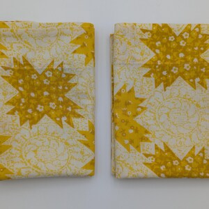 K&ouml;nnte beinhalten: Zwei gefaltete Stoffst&uuml;cke mit einem sich wiederholenden Muster aus gelben Sternen und kleinen wei&szlig;en floralen Akzenten auf cremefarbenem Hintergrund. Der Stoff hat eine Vintage- oder Retro-&Auml;sthetik.