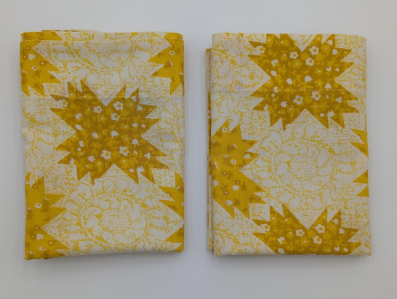 K&ouml;nnte beinhalten: Zwei gefaltete Stoffst&uuml;cke mit einem sich wiederholenden Muster aus gelben Sternen und kleinen wei&szlig;en floralen Akzenten auf cremefarbenem Hintergrund. Der Stoff hat eine Vintage- oder Retro-&Auml;sthetik.