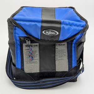 Puede incluir: Bolsa nevera Igloo azul y negra con correa negra para el hombro. La bolsa tiene un bolsillo frontal con cremallera y un bolsillo lateral de malla. El logotipo de Igloo es visible en la parte superior. Para mantener los alimentos frescos.