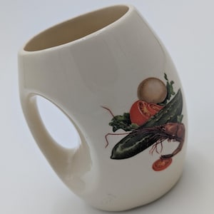 Może przedstawiać: Kremowy ceramiczny kubek o unikalnym kształcie uchwytu. Kubek zdobi kolorowa ilustracja warzyw i raka. Projekt zawiera pomidora, ogórka, pieczarkę i sałatę.