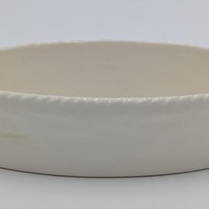Könnte beinhalten: Ovale, cremefarbene Keramikschale mit einem seilartigen Rand. Die Schale hat eine glatte, glänzende Oberfläche und eine geringe Tiefe. Das Design ist schlicht und elegant, geeignet zum Servieren oder Ausstellen.