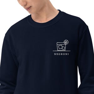 Può includere: Una felpa blu navy a maniche lunghe con un ricamo a linee bianche sul petto sinistro. Il disegno raffigura un bicchiere da cocktail con cubetti di ghiaccio e una fetta di agrume, con la parola NEGRONI stampata al di sotto.