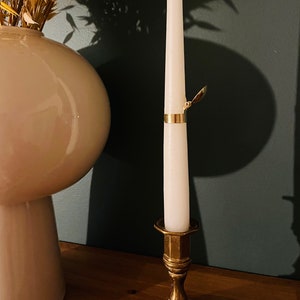 Pode incluir: Uma vela branca cónica num castiçal de metal dourado. A vela tem uma faixa de metal dourado com um desenho de folha. Um vaso branco com flores secas está no fundo.