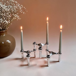 May include: A silver candle holder with three arms and six candle holders. The candle holder is on a light pink surface with three white candles lit.