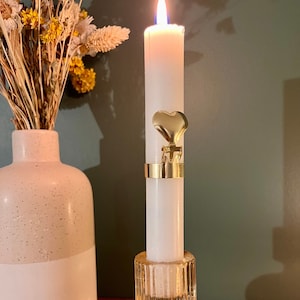 Puede incluir: Una vela blanca con un portavelas dorado en forma de corazón sobre un candelero de vidrio transparente.