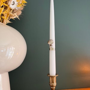 Puede incluir: Una vela blanca con un diseño de concha en el soporte se encuentra sobre un candelabro de latón. El candelabro está sobre una superficie de madera.