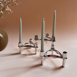 May include: Three silver candle holders with three white candles. The candle holders are triangular in shape with three legs each.
