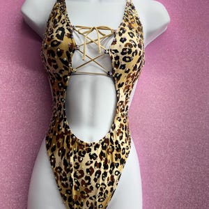 May include: A leopard print swimsuit with a halter neck and a lace-up front. The one-piece swimsuit has a cut-out design and is embellished with rhinestones. The swimsuit is displayed on a white mannequin against a pink background.