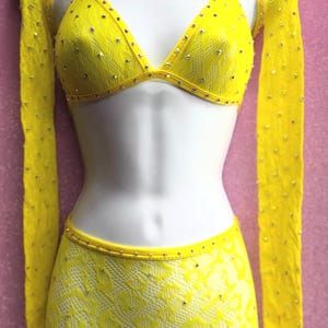 May include: A bright yellow, two-piece outfit with a halter top and shorts, embellished with sparkling rhinestones. The outfit includes long, sheer sleeves and is displayed on a mannequin. The fabric has a delicate, lacy texture.