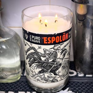 May include: A lit candle in a repurposed glass bottle with a black and white label featuring a skeleton riding a rooster. A bottle of tequila, a cocktail shaker, a knife, and a corkscrew are also visible.