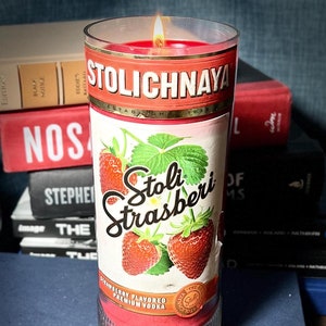 以下が含まれることがあります： 白いラベルに「Stolichnaya Stoli Strasberi Cranberry Flavored Premium Vodka」と書いてある赤いガラスのキャンドル。キャンドルに火が灯され、炎が見えます。