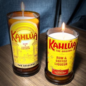 May include: Two glass bottles with yellow labels and black text. The labels read "Kahlua Rum & Coffee Liqueur The Original". One bottle is lit with a candle inside. Both bottles are on a brown wooden surface.