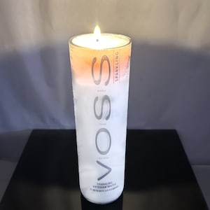 Puede incluir: Una vela encendida dentro de un vaso transparente con el texto "VOSS" y "SPARKLING ARTESIAN WATER" impreso en el lateral. La vela tiene un degradado blanco y naranja, y la llama está encendida.