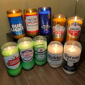 May include: A collection of lit candles in repurposed beer and liquor bottles. The candles feature labels from Bud Light, Budweiser, Heineken, Corona Extra, and other popular brands. The candles are various colors, including yellow, blue, and green.