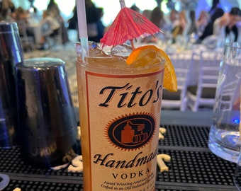 Könnte beinhalten: Ein Cocktail in einem hohen Glas mit dem Etikett Tito's Handmade Vodka. Das Getränk ist mit einer Orangenscheibe und einem kleinen roten Schirm garniert. Ein Strohhalm ist in das Getränk gesteckt. Das Glas steht auf einer Bar.