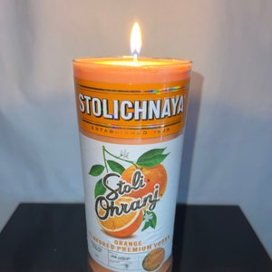 以下が含まれることがあります： オレンジ風味のストリ・オランジ・ウォッカのキャンドル。ガラスの瓶に入っていて、芯に火が灯っています。ラベルにはストリ・オランジのロゴとオレンジのスライスが描かれています。