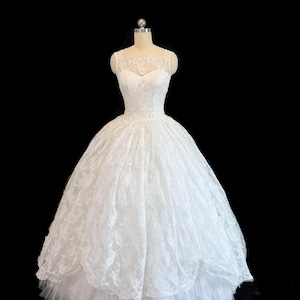 May include: A white floor-length gown featuring a sleeveless bodice with a sheer lace overlay and a sweetheart neckline. The voluminous skirt is crafted from layers of white lace and tulle, creating a full silhouette on a dress form.