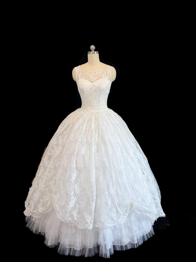 May include: A white floor-length gown featuring a sleeveless bodice with a sheer lace overlay and a sweetheart neckline. The voluminous skirt is crafted from layers of white lace and tulle, creating a full silhouette on a dress form.