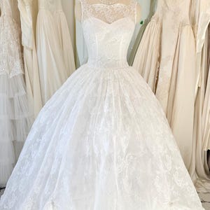 May include: A white floor-length gown featuring a sleeveless bodice with a sheer lace overlay, a sweetheart neckline, and a full, voluminous skirt made of layered lace and tulle fabric. The dress is displayed on a mannequin with a pearl necklace.