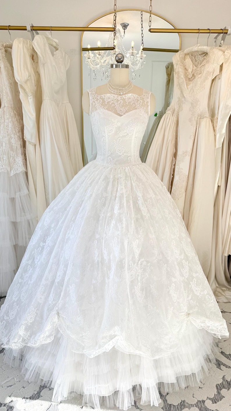 May include: A white floor-length gown featuring a sleeveless bodice with a sheer lace overlay, a sweetheart neckline, and a full, voluminous skirt made of layered lace and tulle fabric. The dress is displayed on a mannequin with a pearl necklace.