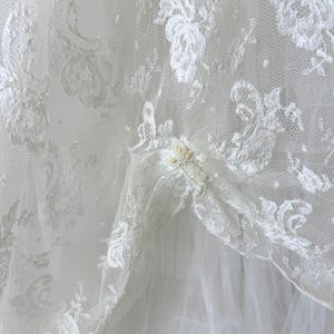 May include: A white lace fabric overlay featuring intricate floral embroidery patterns and delicate beadwork accents. The sheer lace layer is draped over a solid white fabric base, creating a layered texture with visible folds and gathered sections.