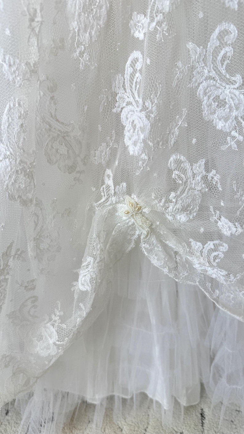 May include: A white lace fabric overlay featuring intricate floral embroidery patterns and delicate beadwork accents. The sheer lace layer is draped over a solid white fabric base, creating a layered texture with visible folds and gathered sections.