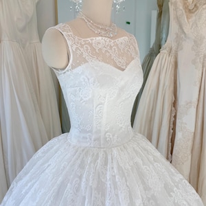 May include: A white sleeveless wedding dress featuring an intricate floral lace overlay on the bodice and a full, voluminous skirt. The gown has a high illusion neckline and a fitted waistline, displayed on a mannequin with a beaded necklace.