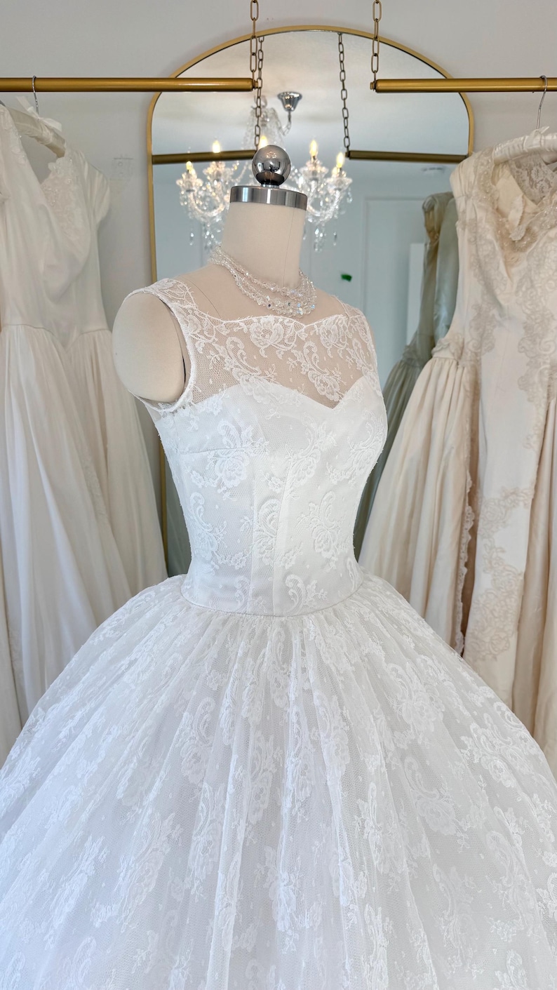 May include: A white sleeveless wedding dress featuring an intricate floral lace overlay on the bodice and a full, voluminous skirt. The gown has a high illusion neckline and a fitted waistline, displayed on a mannequin with a beaded necklace.