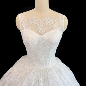 May include: A white sleeveless dress featuring a sweetheart neckline and an overlay of delicate floral lace. The lace extends to the shoulders and upper chest, creating a sheer illusion effect. The bodice is fitted, transitioning into a full skirt.