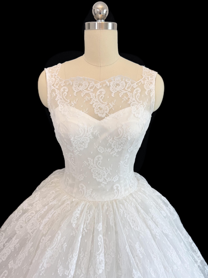 May include: A white sleeveless dress featuring a sweetheart neckline and an overlay of delicate floral lace. The lace extends to the shoulders and upper chest, creating a sheer illusion effect. The bodice is fitted, transitioning into a full skirt.