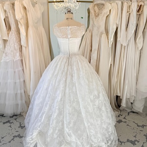 May include: A white floor-length wedding dress on a mannequin, featuring a sheer lace bodice with a square neckline, a fitted waist, and a voluminous skirt with intricate floral lace overlays and a scalloped hemline, secured with a back zipper.