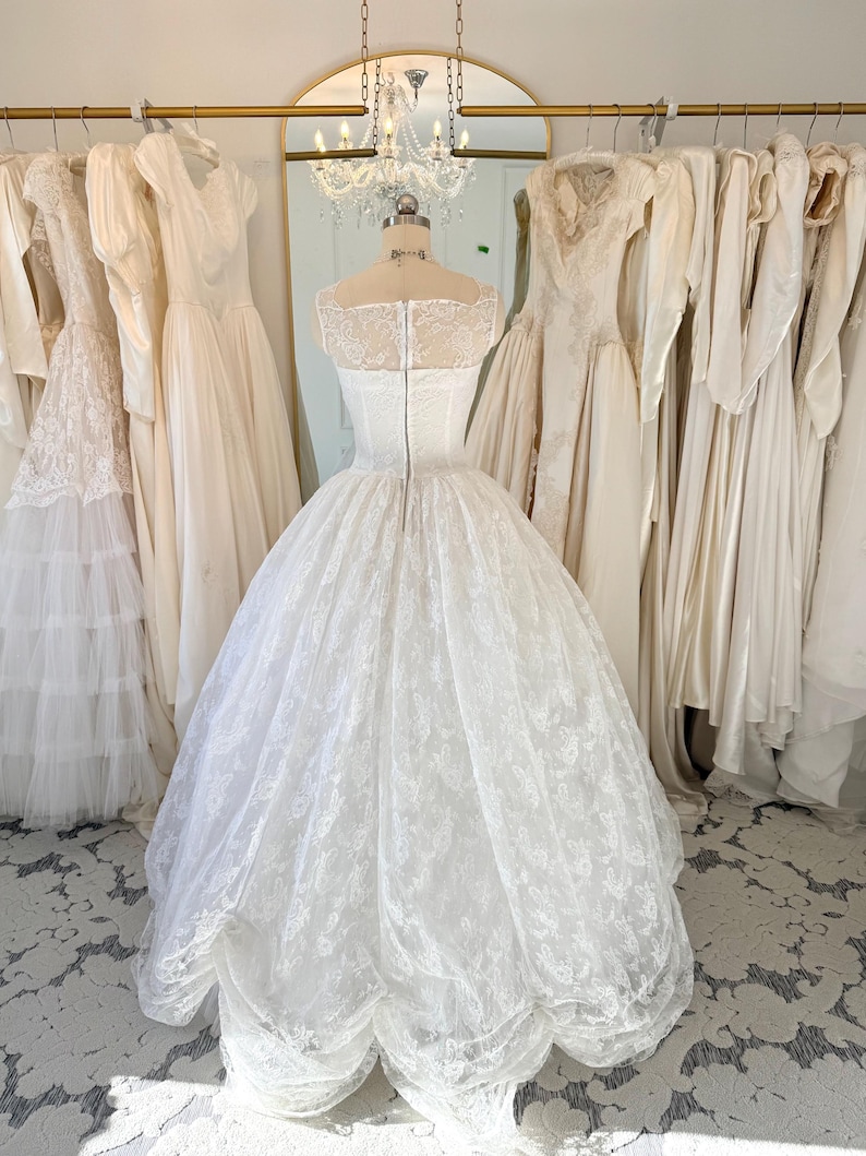 May include: A white floor-length wedding dress on a mannequin, featuring a sheer lace bodice with a square neckline, a fitted waist, and a voluminous skirt with intricate floral lace overlays and a scalloped hemline, secured with a back zipper.