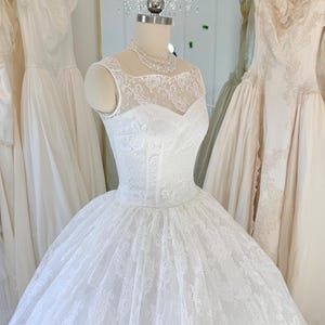 May include: A white sleeveless gown featuring a fitted bodice with a sweetheart neckline and a sheer lace overlay. The dress has a full, voluminous skirt covered in intricate floral lace patterns, displayed on a mannequin with a pearl necklace.
