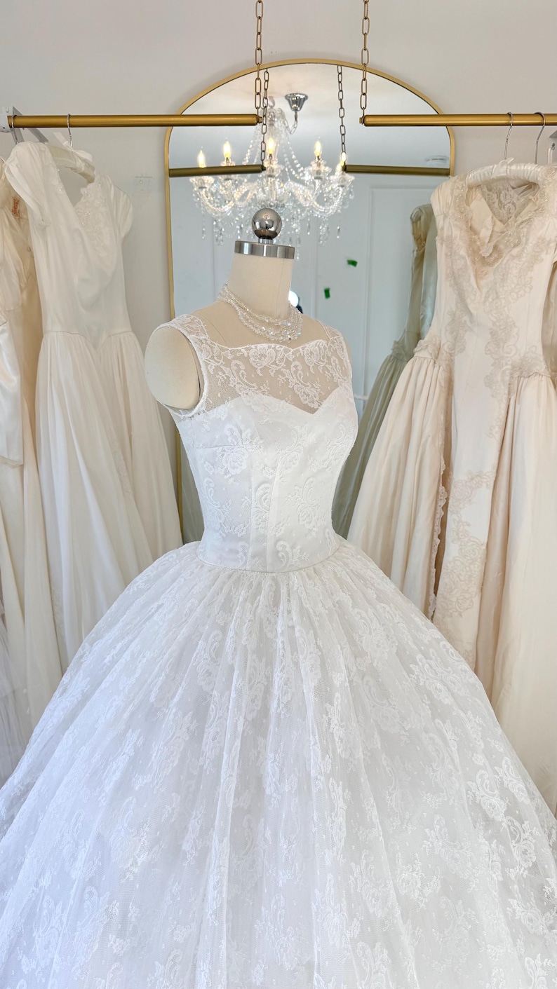 May include: A white sleeveless gown featuring a fitted bodice with a sweetheart neckline and a sheer lace overlay. The dress has a full, voluminous skirt covered in intricate floral lace patterns, displayed on a mannequin with a pearl necklace.