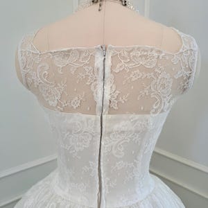 May include: A white lace dress featuring a sheer lace bodice with a floral pattern, a square neckline, and a vertical center back zipper closure. The garment is displayed on a mannequin and is paired with a multi-strand beaded necklace.