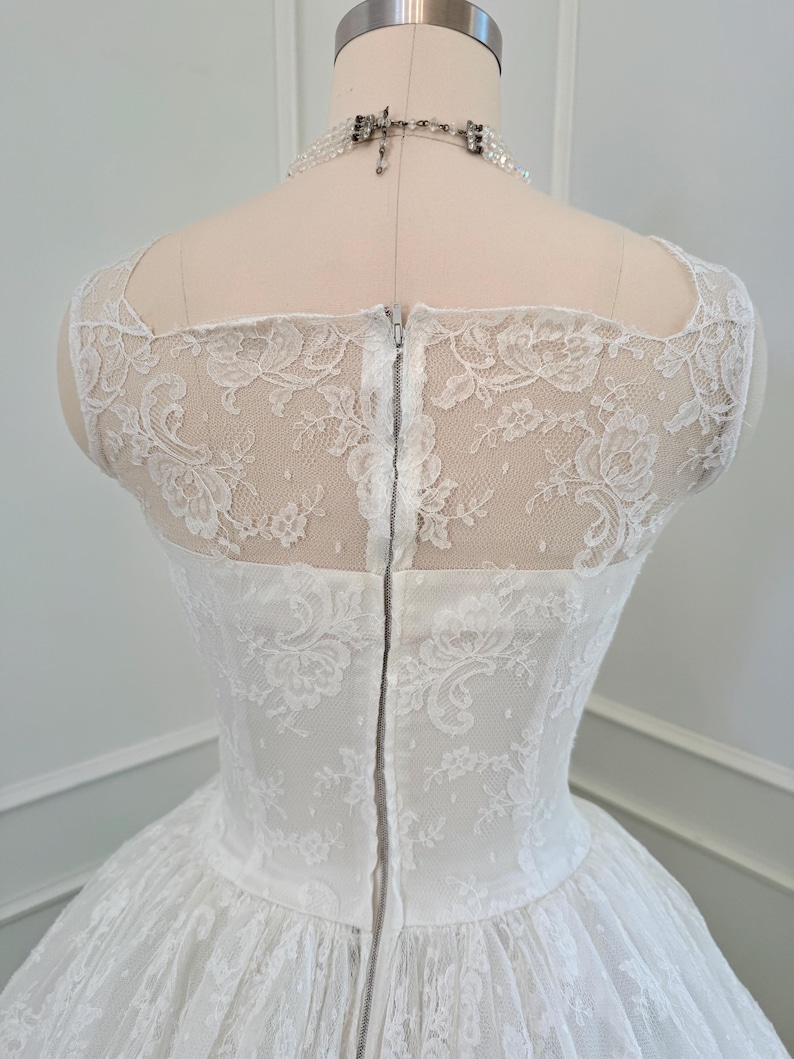 May include: A white lace dress featuring a sheer lace bodice with a floral pattern, a square neckline, and a vertical center back zipper closure. The garment is displayed on a mannequin and is paired with a multi-strand beaded necklace.