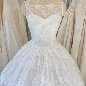 May include: A sleeveless white wedding dress featuring a sheer lace overlay with intricate floral patterns and delicate beadwork. The bodice has a sweetheart neckline under a high illusion lace yoke, transitioning into a full, voluminous skirt.